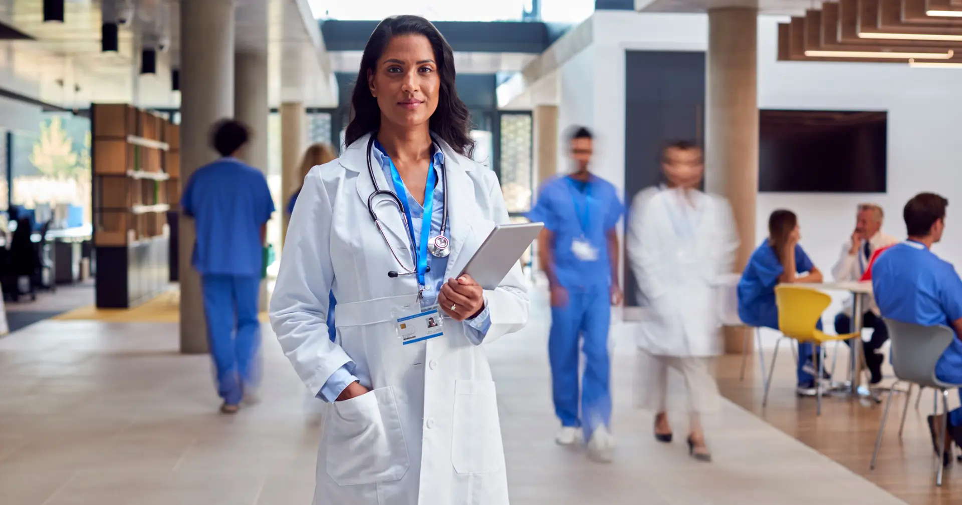 Female Doctor Wearing White Coat With Digital Tablet In Busy Hospital